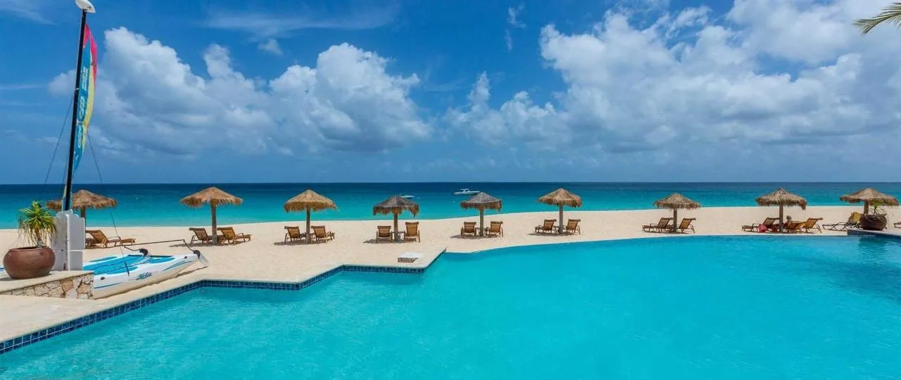 A resort swimming pool borders a sandy beach lined with thatched umbrellas and lounge chairs overlooking turquoise waters in Anguilla.