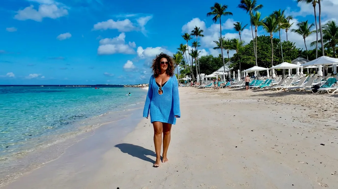 A woman in a blue knit cover-up walks barefoot along a white-sand beach lined with palm trees in Punta Cana.