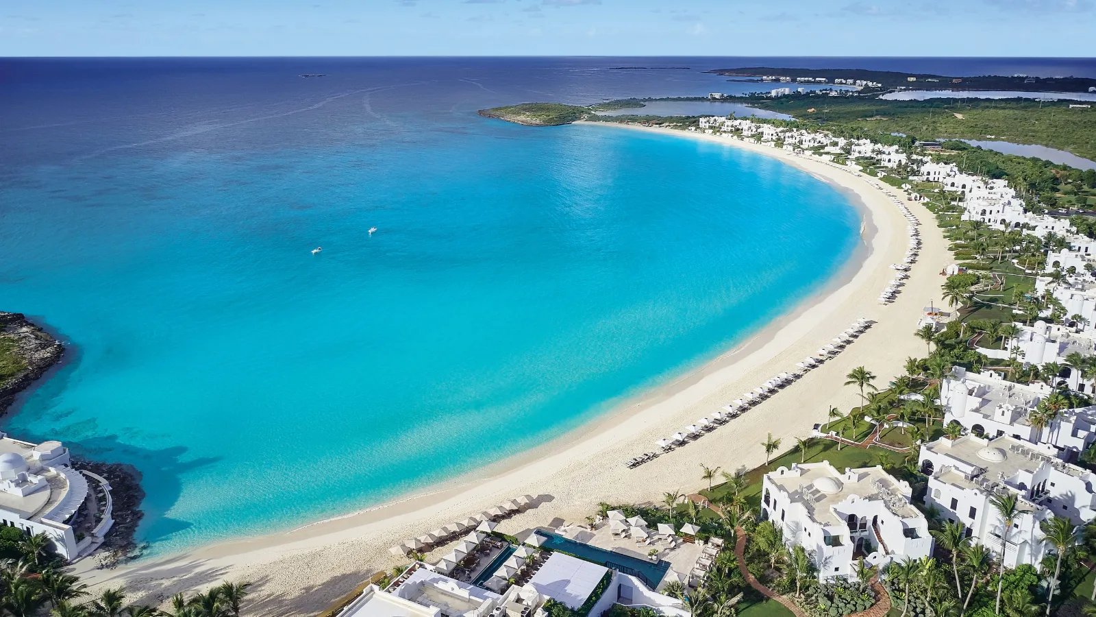 White resort villas line a crescent-shaped white sand beach and turquoise bay along the coast of Anguilla.