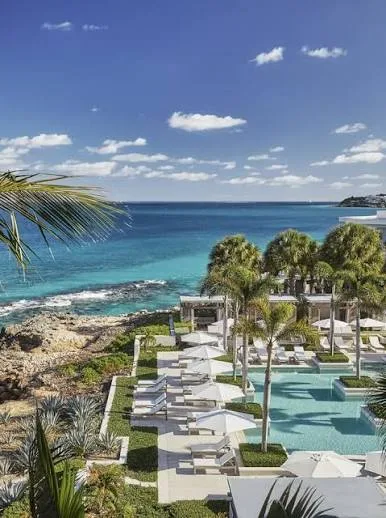 White sun loungers and umbrellas line a resort pool overlooking the turquoise sea and rocky coast in Anguilla.