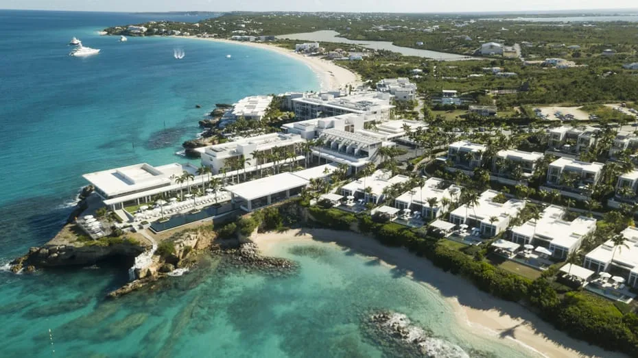 Modern white resort buildings and villas line a rocky coastline overlooking turquoise ocean waters and sandy beaches in Anguilla.