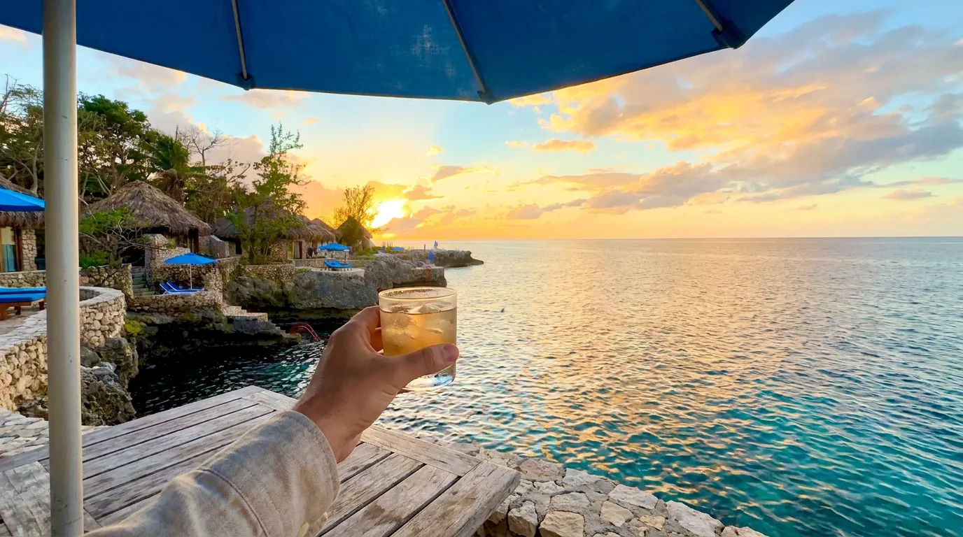 A hand holds a cocktail under an umbrella on a cliffside terrace overlooking thatched villas and a sunset in Jamaica.